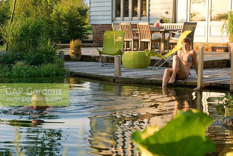 Couple enjoying swimming pond on warm summer's evening with decking and seating in background - Bressingham, Norfolk.