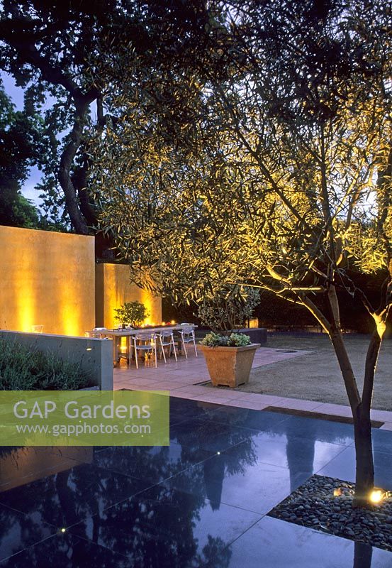 Modern garden with black stone paving, tree and dining area lit up in the evening - California, USA
