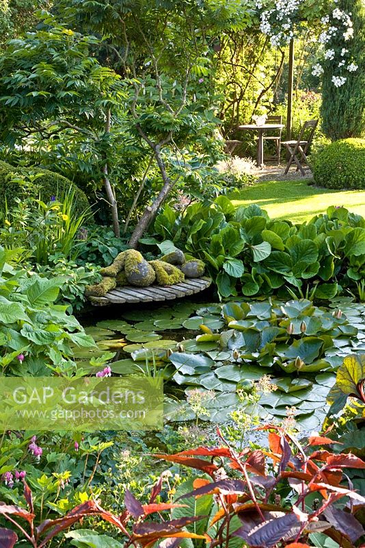 Pond in country garden with reclining sculpture on wooden decking and seating area in background