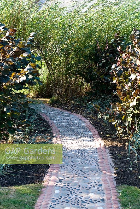 Path with a daisy design made from pebbles and stones set in coloured concrete leading to a willow tunnel with Corylus avelana 'Purpurea' and grasses on either side