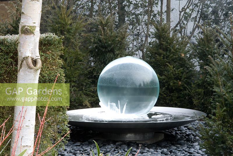 Glass ball fountain with icicles at the base with running water, black pebbles and a circular yew hedge - Betula utilis jacquemontii and Cornus sanguinea 'Winter Beauty' in foreground