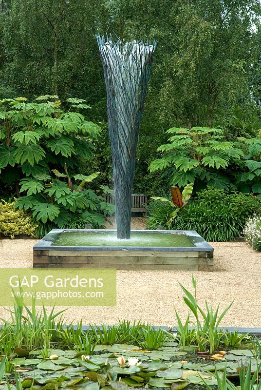 Fountain in scultured ironwork with wooden seat and tetrapanax papyriferus growing on either side - The Exotic Garden, The Old Vicarage, East Ruston, Norfolk