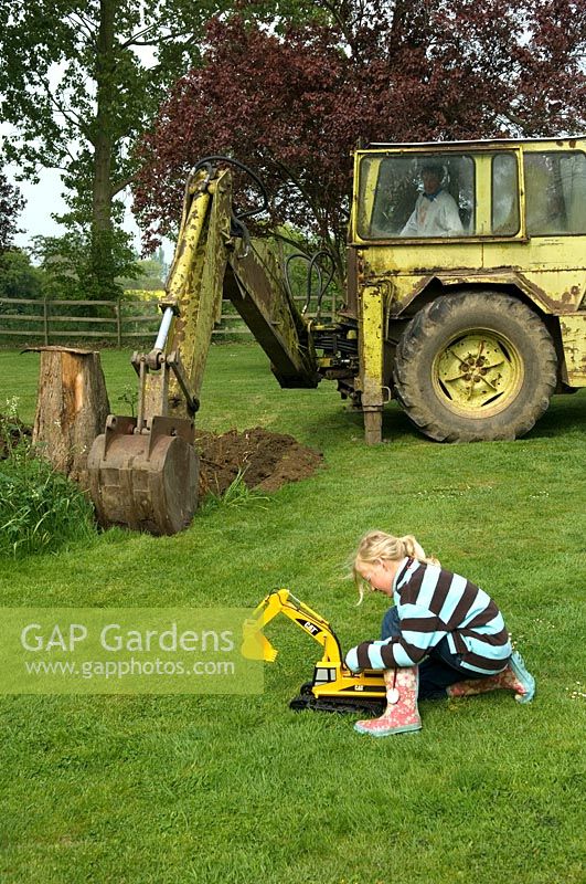 Child playing with toy digger on lawn with real digger digging hole behind - Pannells Ash Farm, West Essex