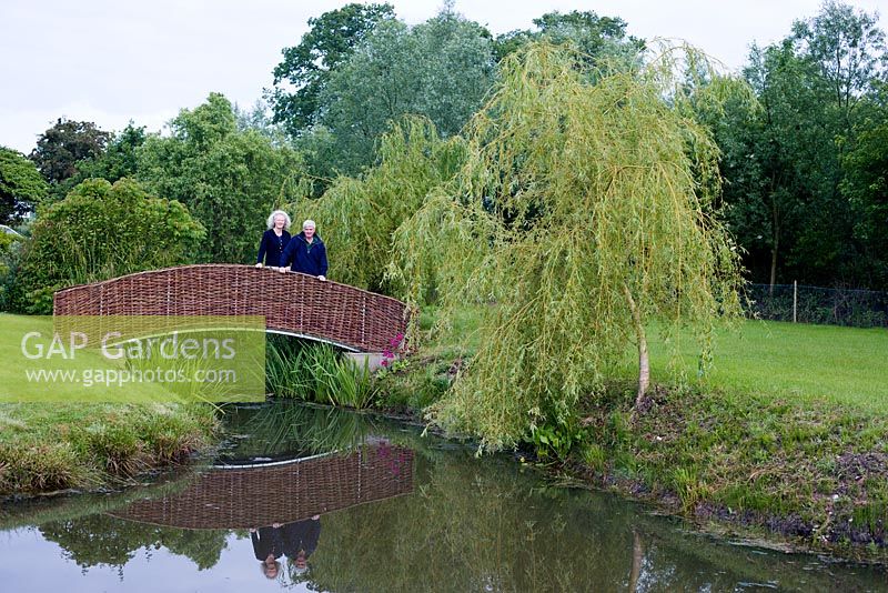 Susie and Robert Yates on willow bridge across pond with Salix x sepulcralis var. chrysocoma and candelabra primulas to right.