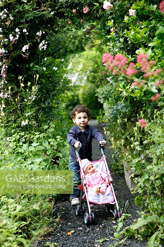 Small girl pushing doll in pink pram along a gravel stone garden path, under an arch of Roses