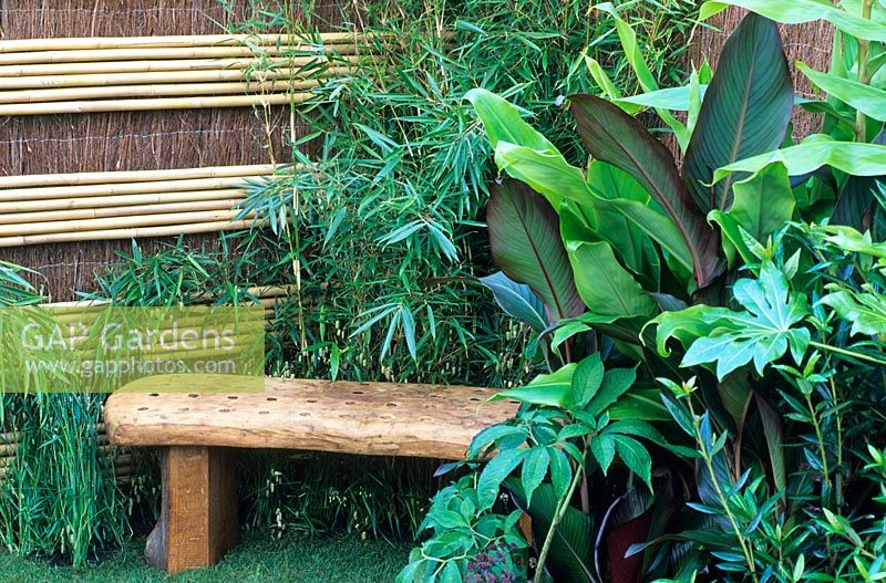 Foliage plants surround a wooden bench with a bamboo and brushwood screen fence in the 'Hideaway' garden, RHS Hampton Court Flower Show
