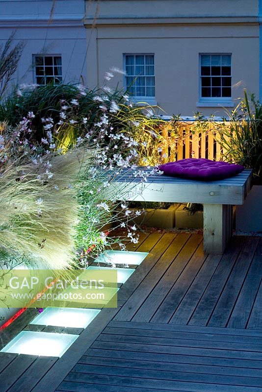 Decked terrace at night with pink and white led lighting and wooden bench - Roof garden, Holland Park, London