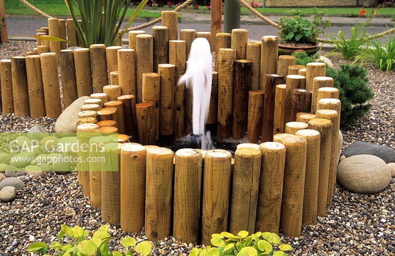 Sunken small water feature with fountain surrounded by lengths of short wooden stakes, gravel and pebbles 