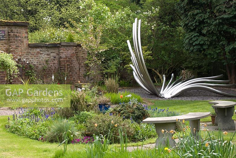 Curved stone benches overlooking small cicular bed in Dave's Garden and 'Nature's Breeze', a stainless steel tubular sculpture created by Sue Sharples, inspired by studies of yucca and cordyline plants - Poulton Hall, NGS garden, Cheshire