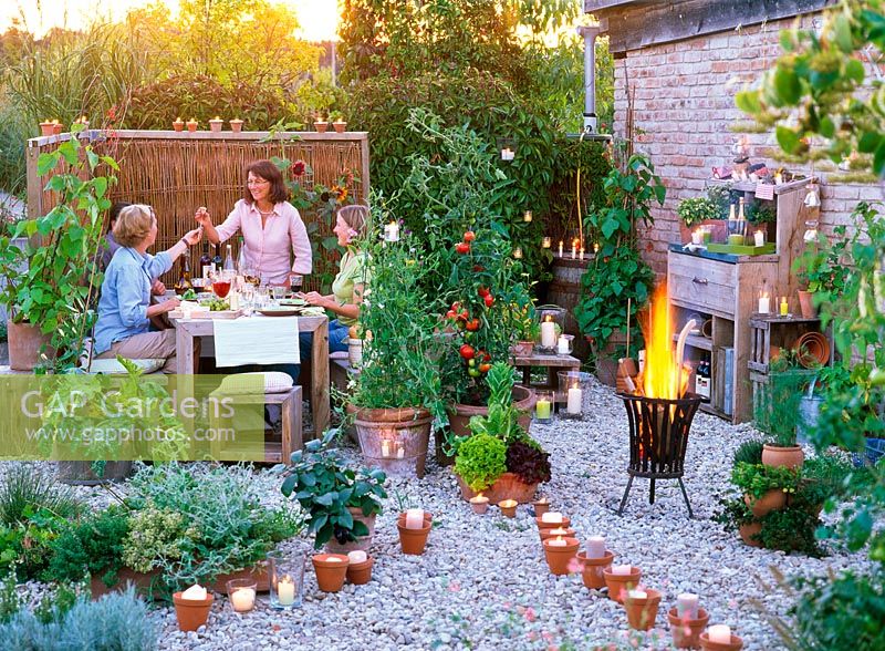 People using dining area, gravel pathway lit with candles, container plants include vegetables and herbs