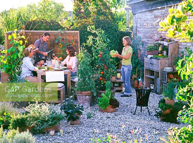 People using dining area, gravel pathway lit with candles, container plants include vegetables and herbs