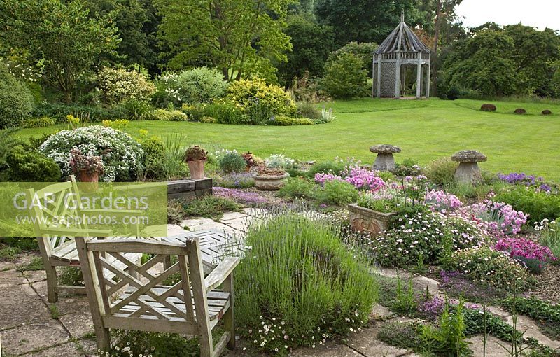 Overall view of alpine garden planted with many aromatic alpines with wooden chairs looking out onto lawns with borders of herbaceous perennials and beautiful green oak gazebo - Woodpeckers, Warwickshire