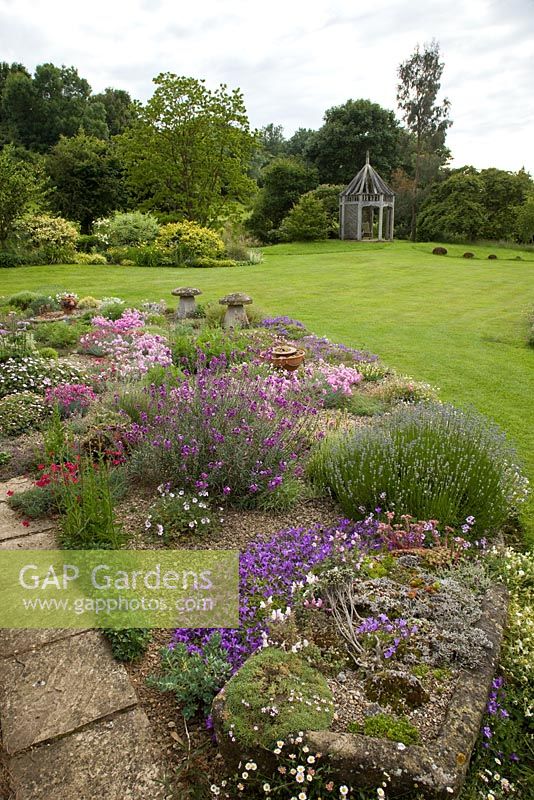 Garden view of alpine garden planted with many aromatic alpines planted in gravel old stone troughs looking out onto lawns and borders of herbaceous perennials - Woodpeckers, Warwickshire