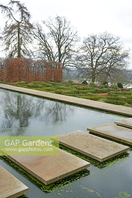 Walkway of concrete paving stones across pond, hornbeam hedge behind and box parterre - Broughton Grange in February 