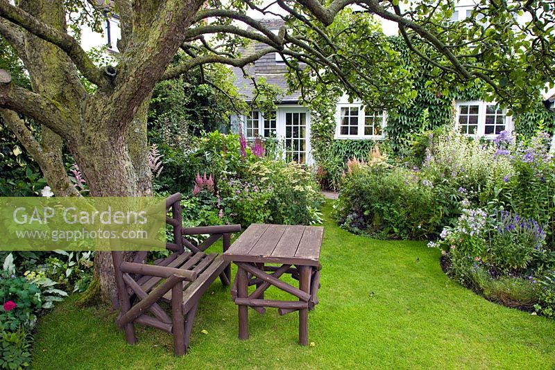 Borders of herbaceous perennials rustic bench and table under old tree with flowers in profusion, packed into an idyllic English cottage garden, at Grafton Cottage ,NGS, Barton-under-Needwood Staffordshire
