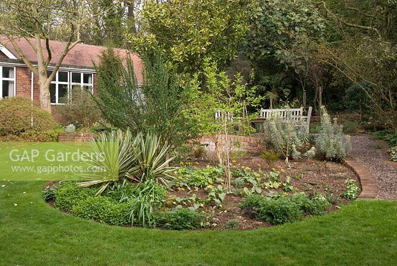 Spring view of secluded garden with mature shrubs, circular central bed, brick wall and gravel path leading to seating area at the back of the bungalow at 'Briarfield' Cheshire