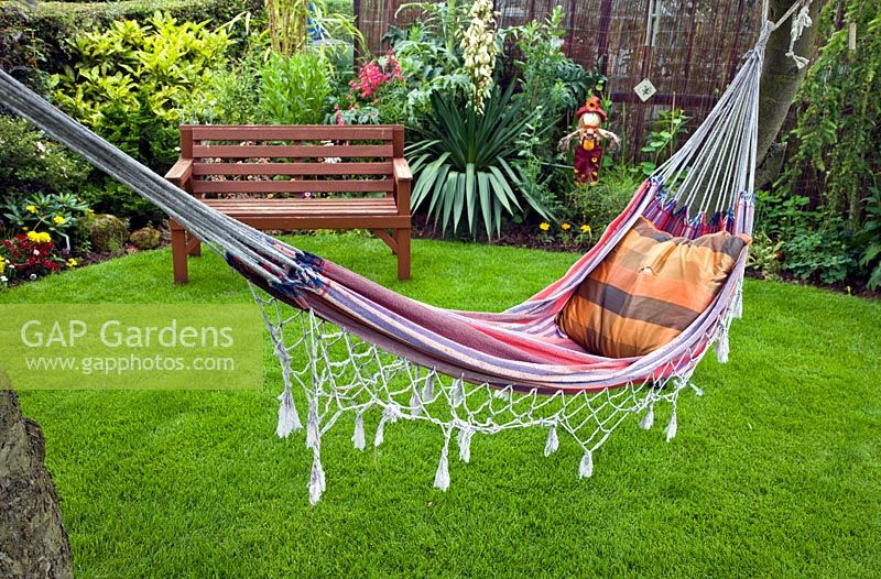 Colourful stripped hammock suspended from trees border of mixed herbaceous perennials wooden bench on lawn