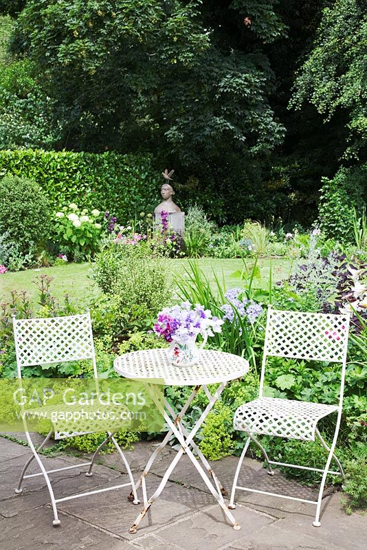 Small urban garden packed full of plants simply designed around a central circular lawn. Lattice work iron chairs and small table with Sweet Peas in jug. York stone patio with border and lawn behind. Sculpture by Christopher Marvell. Alchemilla mollis, Campanula lactiflora, Cotinus, Pittosporum tenuifolium.