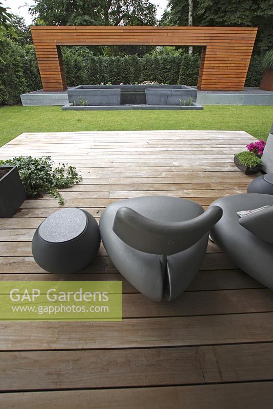 Terrace in modern garden with designer chairs on wooden decking. Artistic wooden arch with raised pond.