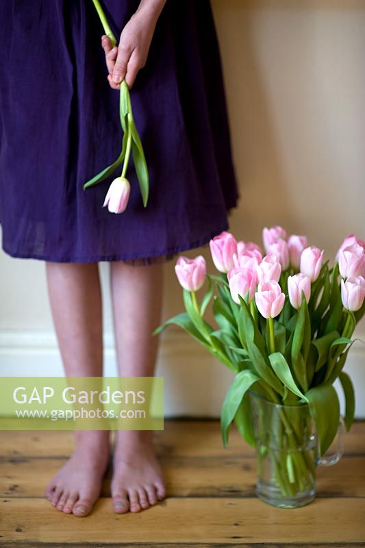 Woman in a dress, standing barefoot alongside a bunch of pink tulips in a vase 