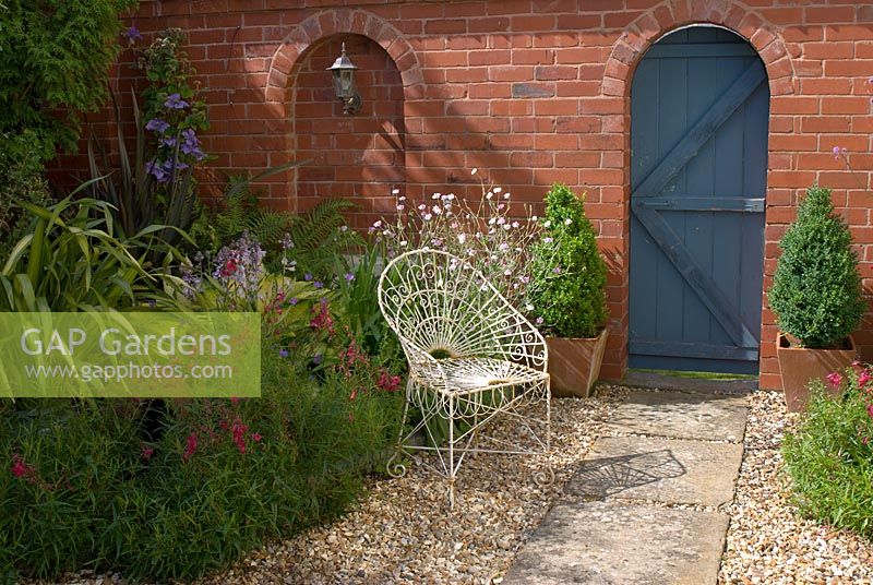 Timber garden gate in brick wall. Stone slab and gravel path with wirework chair. 'Hazelwood'. Jacqueline Iddon Hardy Plants, NGS garden, Lancashire 