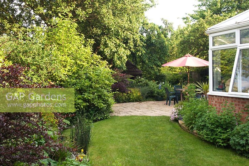 Patio in cottage garden with parasol over seating area. Windy Corner NGS, Barton, Cambs, UK 