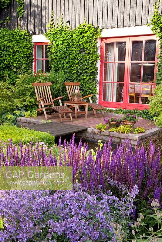 Patio in country garden with wooden loungers. Borders of Nepeta nervosa and Salvia nemorosa 'Caradonna'