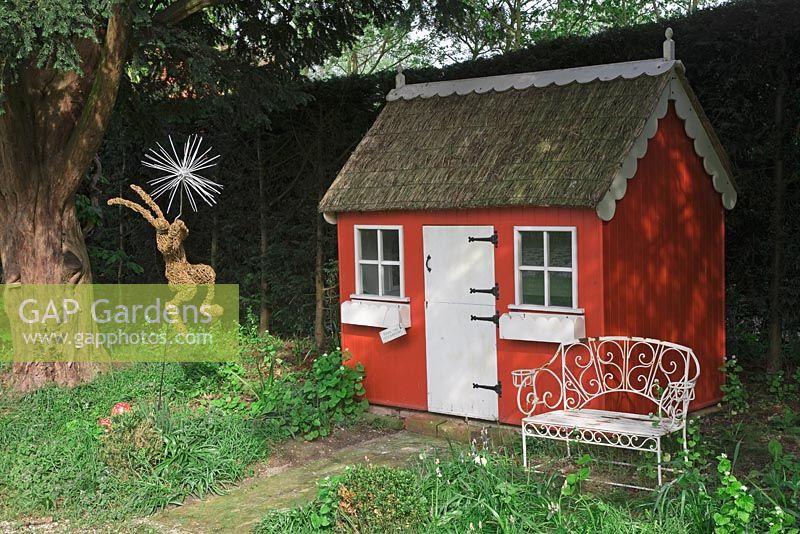 Children's playhouse with thatched roof, wrought iron bench and willow woven hare under shady tree - Northend