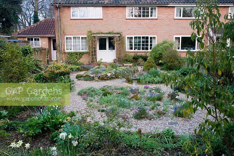 View of alpine front garden in spring with sundial. Plants include Narcissus, Pulsatilla vulgaris, Skimmia japonica, dwarf conifer Picea abies 'Gregoryana', various species of Muscari - Grape Hyacinth, Tulipa, Primula seedlings and Euphorbia characias 'Portuguese Velvet'.