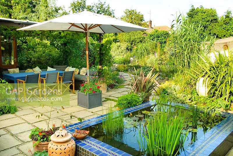 View across raised pool and terrace with covered outdoor seating under pergola clad with Vitis - Grape vine. Canvas parasol with flower filled planter base.