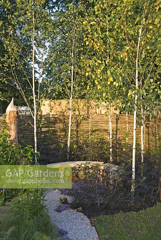 Cockleshell path leading to a seat stacked with logs underneath for wildlife, surrounded by Willow fencing, Betula utilis var. jacquemonii and Sambucus nigra 'Black Lace'. 'It's Only Natural' - Silver Gilt Medal Winner - RHS Hampton Court Flower Show 2010 
 