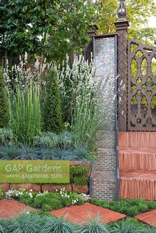 Rainbow effect water feature next to herringbone tile steps, leading up to a carved wooden gate.  Plants include Lavandula angustifolia 'Alba' Koeleria glauca, Armeria maritima 'Alba' and clipped Ligustrum. 'The Garden Lounge' - Silver Gilt Medal Winner - RHS Hampton Court Flower Show 2010 
 