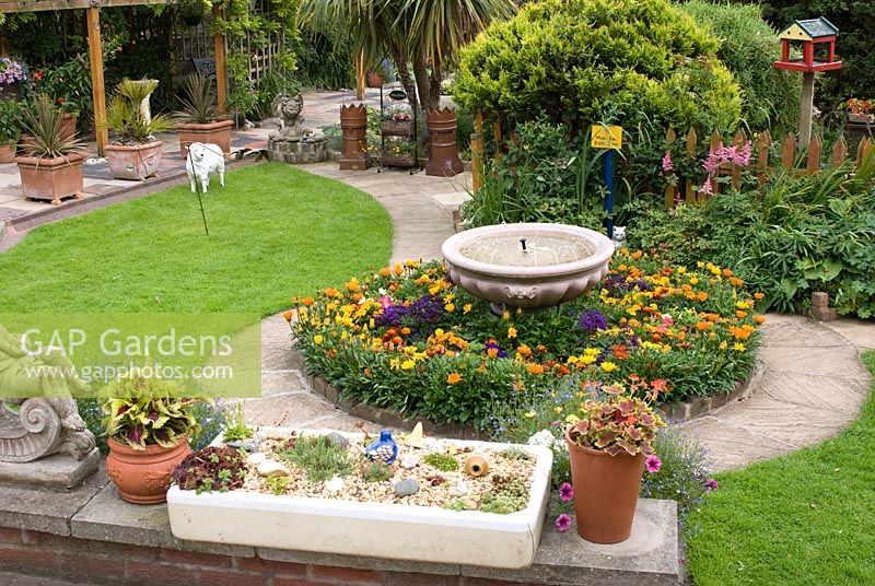 Back garden wall with old stone sink overlooking lawn, water feature with bedding plants, and curved path and trellis walkway with a variety of interesting reclaimed items. Meadow Ave, Southport, Lancashire. The garden opens for The National Gardens Scheme