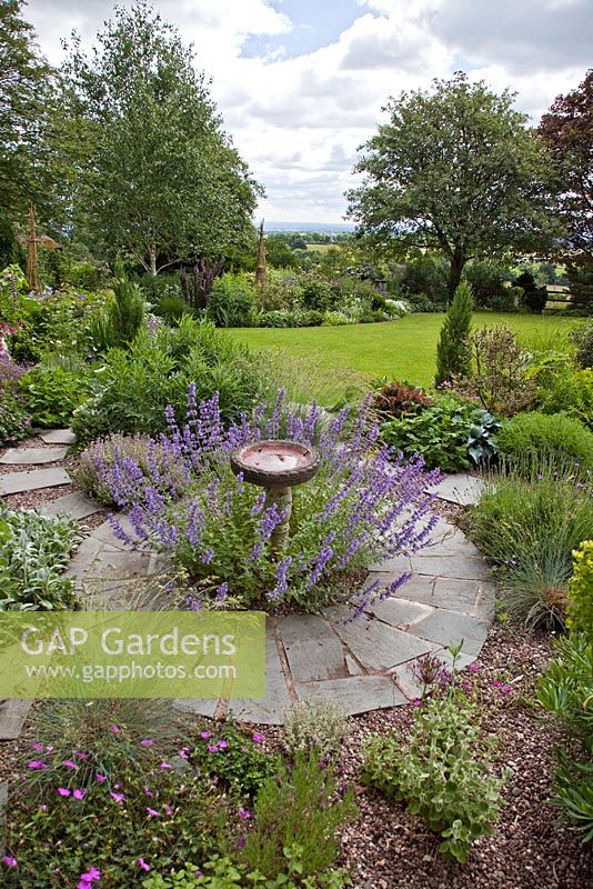Borders of herbaceous perennials, circular slate area with ornamental bird bath underplanted with Nepeta racemosa 'Walkers Low'. Lilac Cottage (NGS) Garden, Gentleshaw, Staffordshire, UK 
 