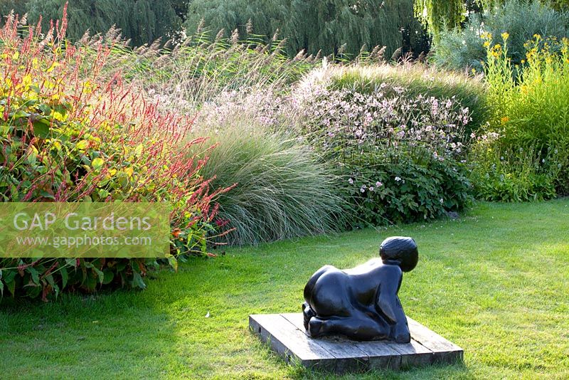 Crouching figure sculpture on lawn and border of Anemone 'Herbstanemone', Festuca 'Marei', Persicaria amplexicaulis 'Firetail', Anemone tomentosa 'Robustissima'