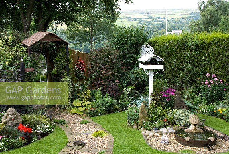 Compact corner of small back garden overlooking hills in distance with mixed borders, Leylandii hedge, gravel and cobble path with mat forming plants leading to ornate gate in wooden fence, and features including - a painted bird table, a half barrel water feature with submerged head waterspout and hands, stone gargoyle, clock face made from painted table top and carved stone sculpture with 'Croeso' - Welcome in Welsh at 'Trevinia', Stubbins, Lancashire NGS
 