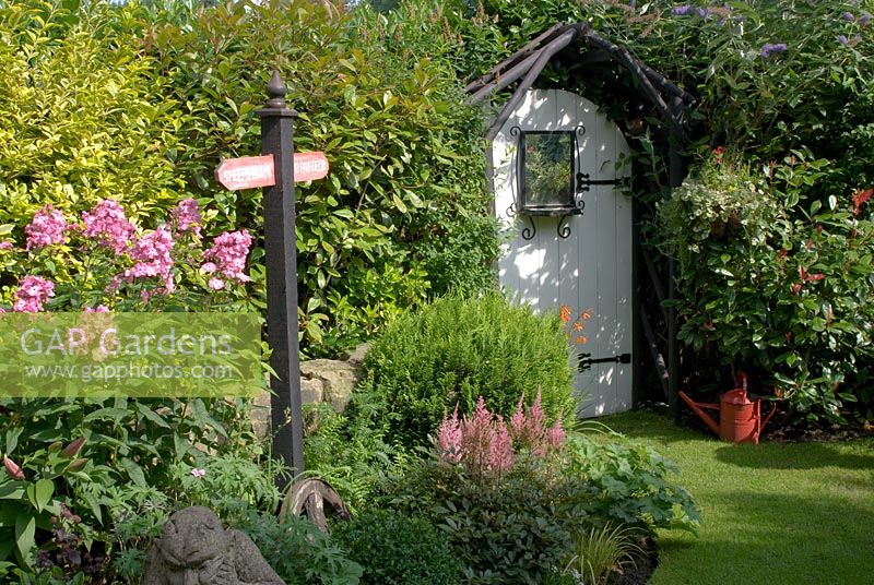 Small back garden with mixed planting including Phlox, Astilbe, conifer, Elaeagnus, Photinia 'Red Robin', Buddleia, and features of signpost, painted watering can and wooden painted arched gate with ornate mirror - 'Trevinia', Stubbins, Lancashire, NGS
