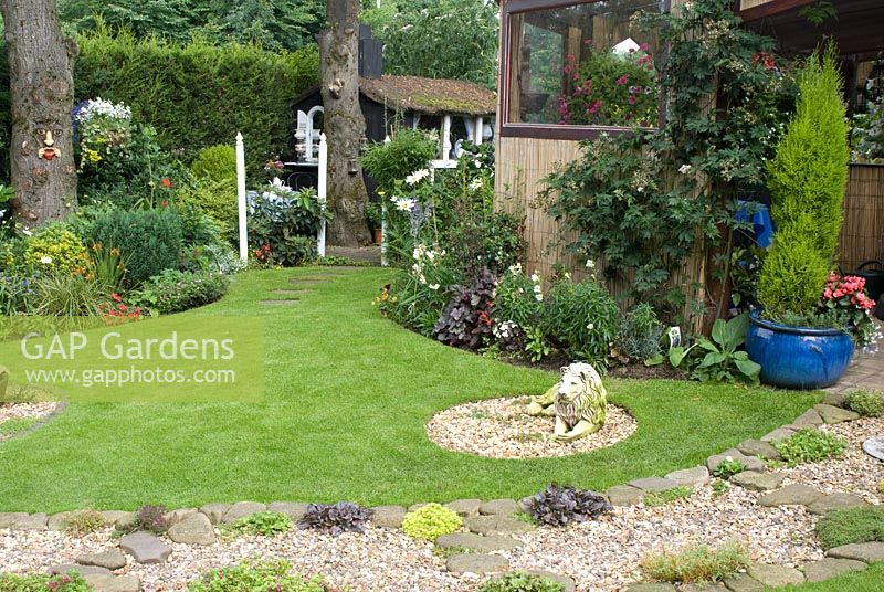 Back garden with lawn and stone lion statue, border with trained Rubus fruticosus, gravel and cobble path with mat forming plants leading to covered outdoor living area with bamboo screens and containers - 'Trevinia', Stubbins, Lancashire, NGS