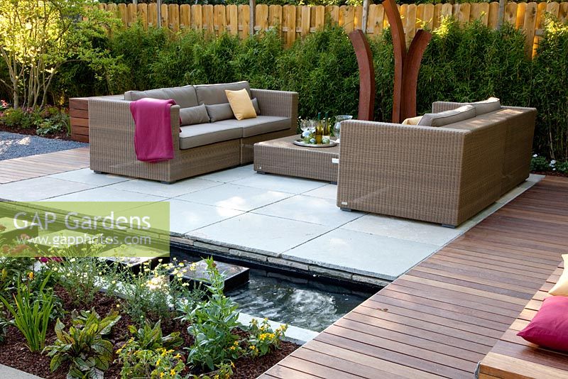 Small garden with wicker sofas on decked and paved patio, backed by Fargesia murielae - Bamboo hedge
 
