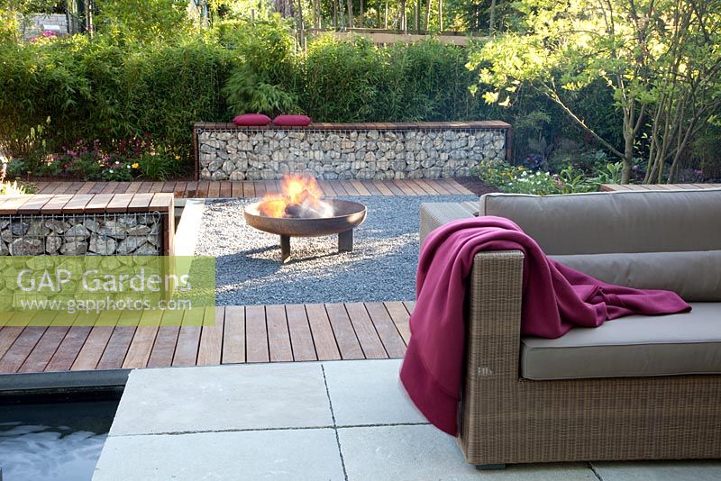 Small garden with wicker sofas and Gabion benches on decked and paved patio, backed by Fargesia murielae - Bamboo hedge. Square gravel area with fire pit