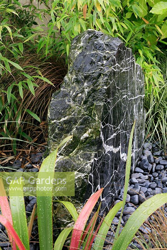 Drilled marbled rock discharging water through a bed of pebbles into an underground sump and surrounded with grassy leaved foliage including bamboo, Phormium and Carex