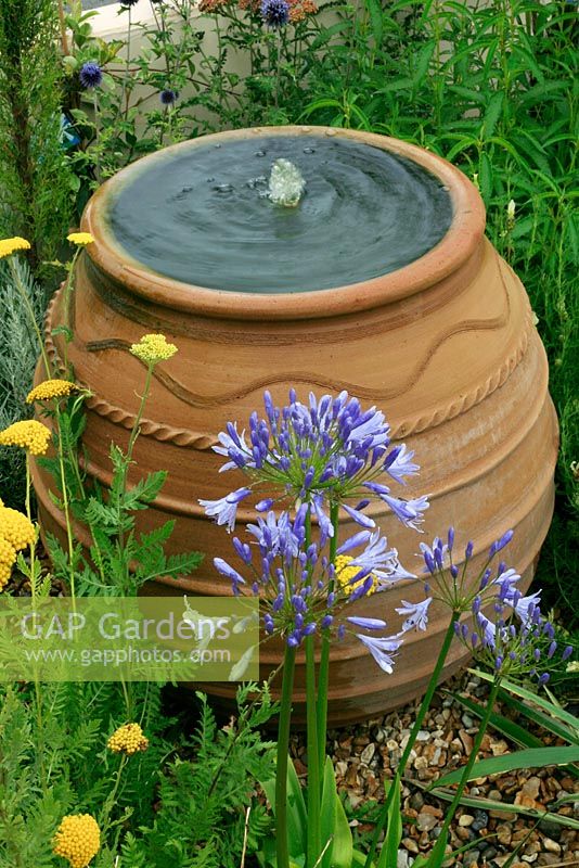 Cretan terracotta oil jar fountain discharging water into a gravel bed with Agapanthus and Achillea in the foreground