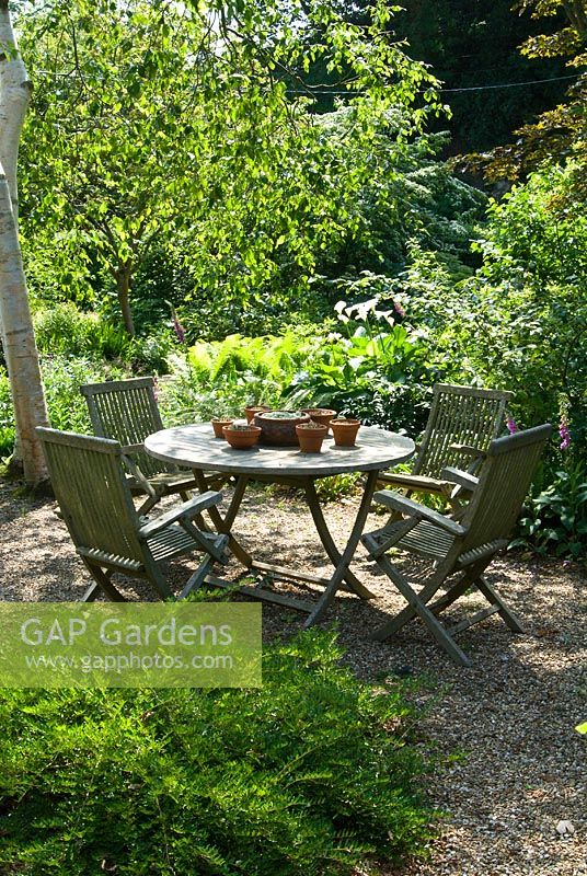 Wooden table and chairs in front woodland garden, June. Sandhill Farm House, Hampshire.