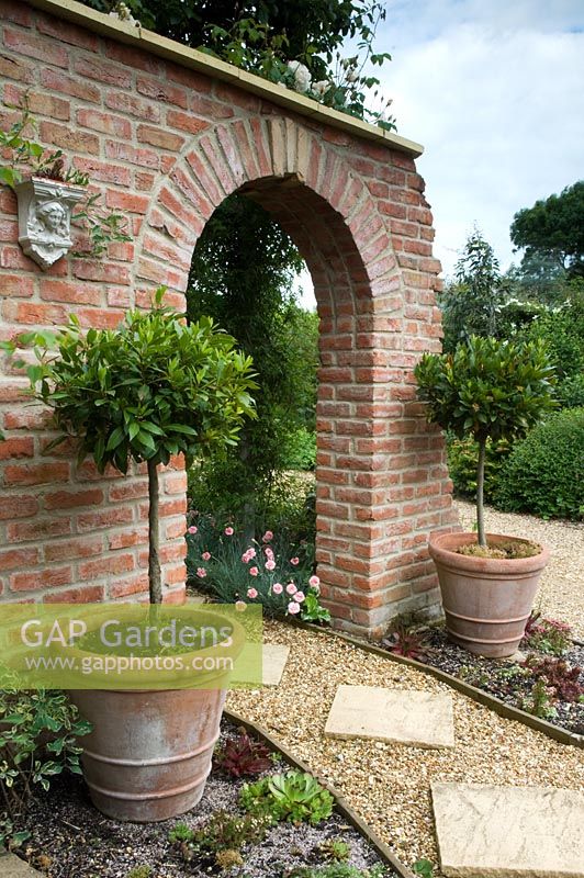 Brick wall with arch and pots with standard Laurus nobilis - Bay trees. Dianthus edging diagonal stepping stone path. Parsons Cottage