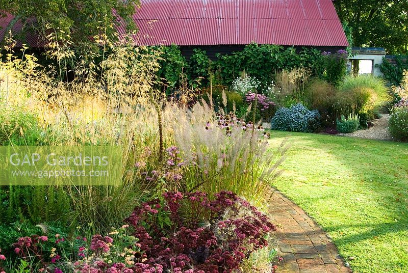 Sedum 'Purple Emperor' in foreground echoes colour of tin roof beyond, surrounded by Stipa gigantea, fluffy Calamagrostis brachytricha and Achillea - Grass Garden, Hants
