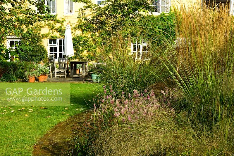 Central circular bed is a mass of graceful grasses and herbaceous perennials including Miscanthus 'Morning Light', Stipa gigantea, Calamagrostis x acutiflora 'Karl Foerster', Allium pulchellum and bronze fennel with house behind - Grass Garden, Hants