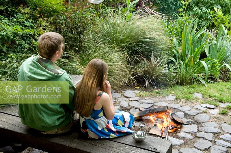 Man and child sitting on wooden bench by firepit on circular cobblestone patio in urban garden. Yulia Badian, London, UK