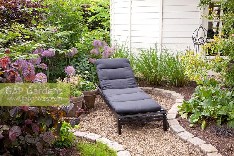 Small gravel patio with lounger surrounded by planting of Corylus avellana 'Purpurea' and Alliums