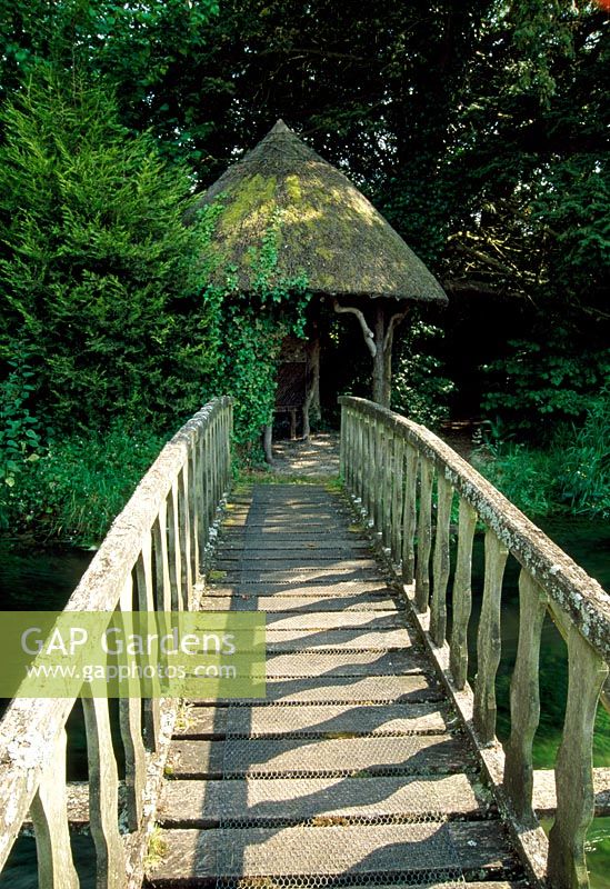 Wooden bridge over river leading to rustic thatched summerhouse - Weir House, Hants