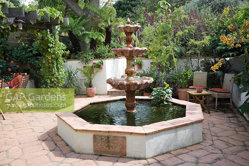 Marble Italianite tiered fountain in octagonal raised pool on terracotta tiles. Vine on oak pergola and wicker chairs in small urban garden